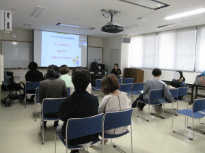 小児科医師による講演の様子。プロジェクターで奥のスクリーンに講座の内容を投影している。人々が椅子に座ってきいている。
